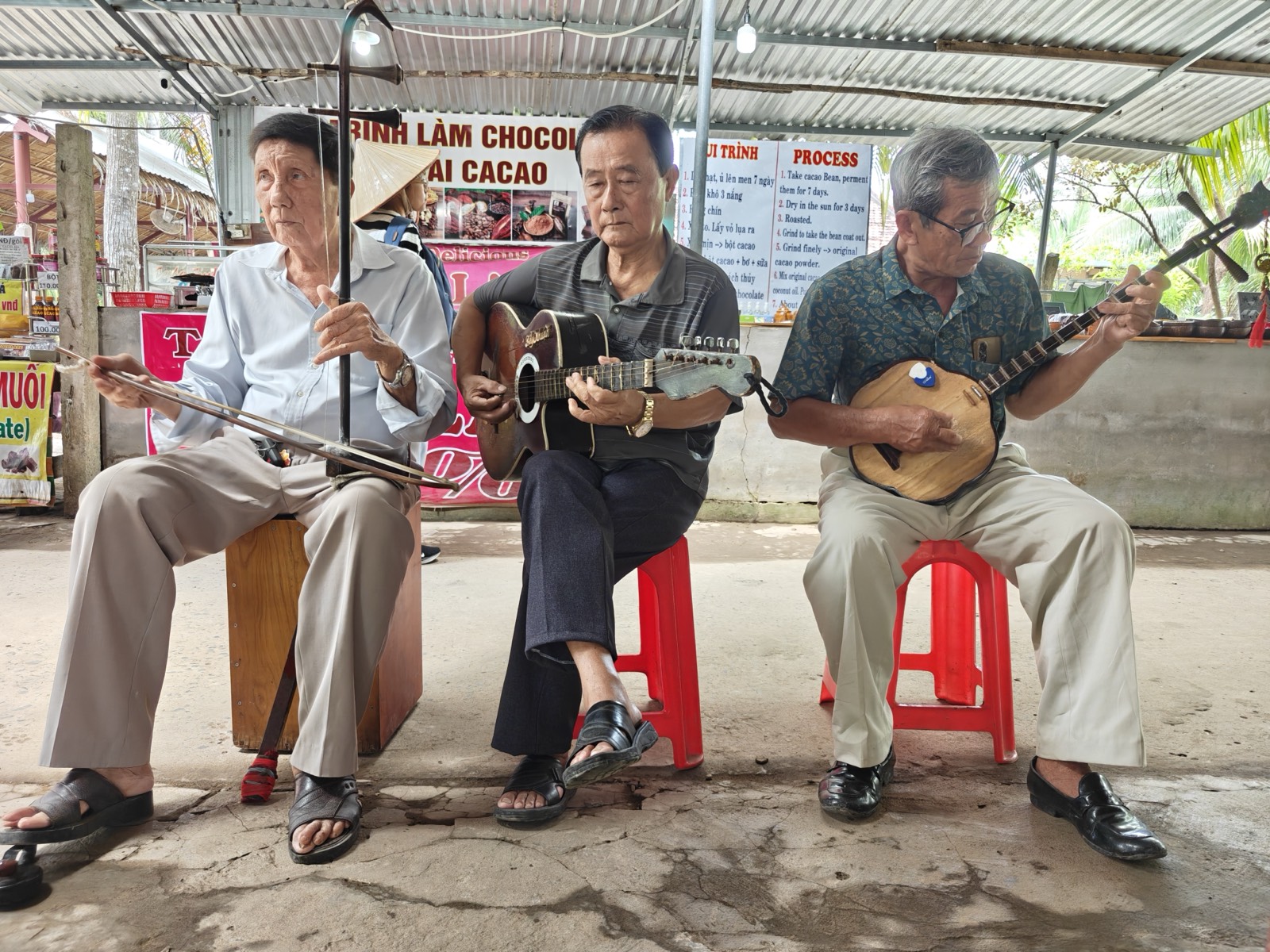 Three men seated under a corrugated roof play a bowed two-string fiddle, a guitar and a round-bodied lute; posters and palm trees behind them.