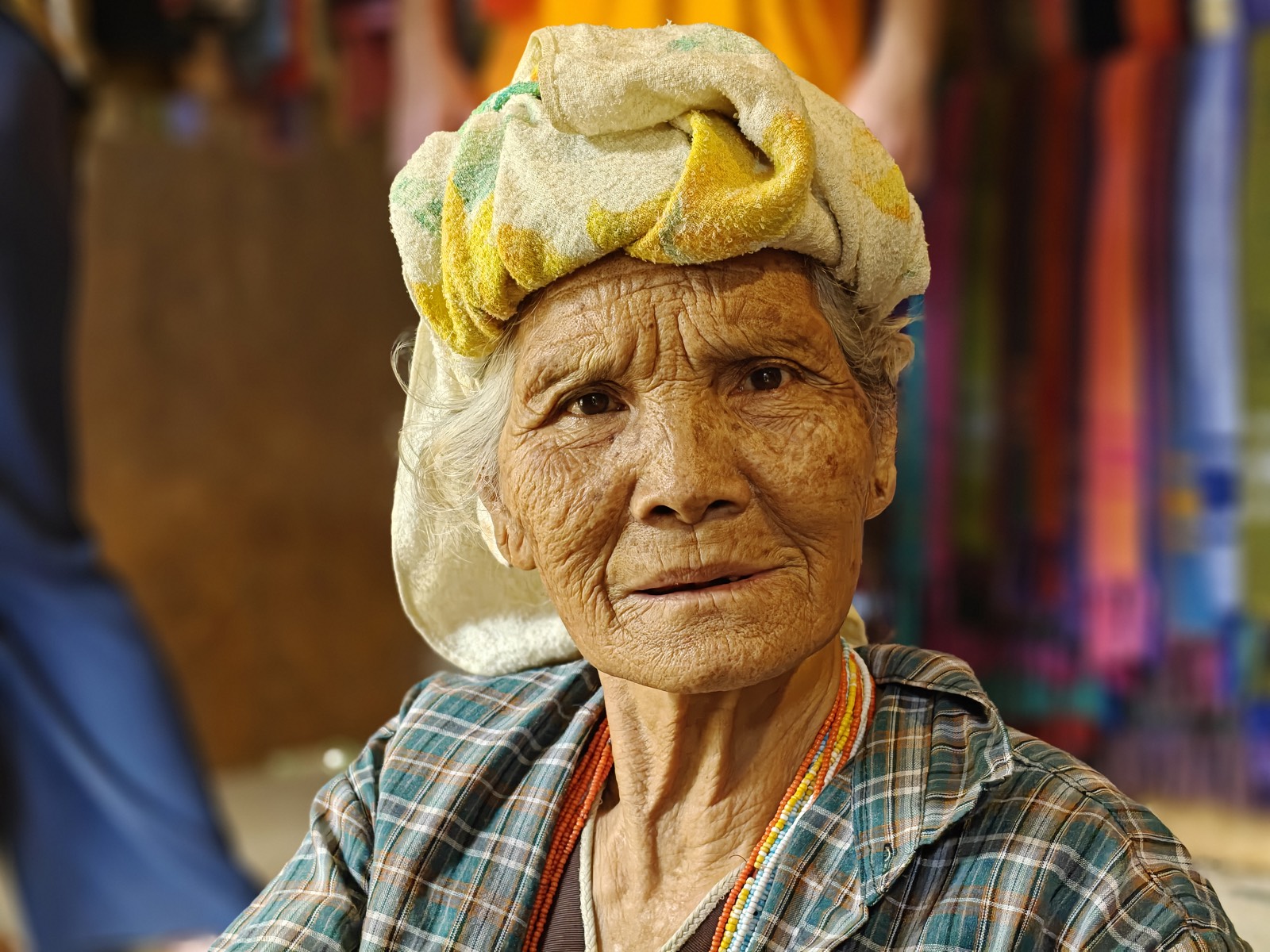 An elderly Karen weaver looks straight at the camera, a towel wrapped on her head, orange and white beaded necklaces over a plaid shirt; woven fabrics hang behind her.